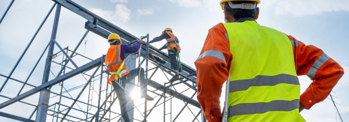 A man in a hi-vis jacket observes two construction workers on a building frame