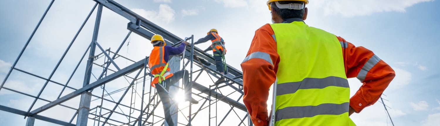 A man in a hi-vis jacket observes two construction workers on a building frame