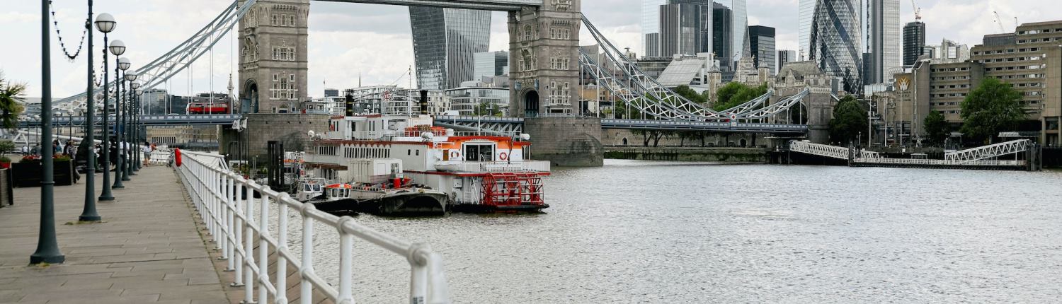 Boats moored alongside the banks of the River Thames in London, with Tower Bridge and skyscrapers in the City of London in the background