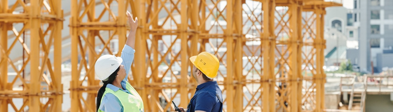 A woman and a man wearing hard hats look toward a steel building structure